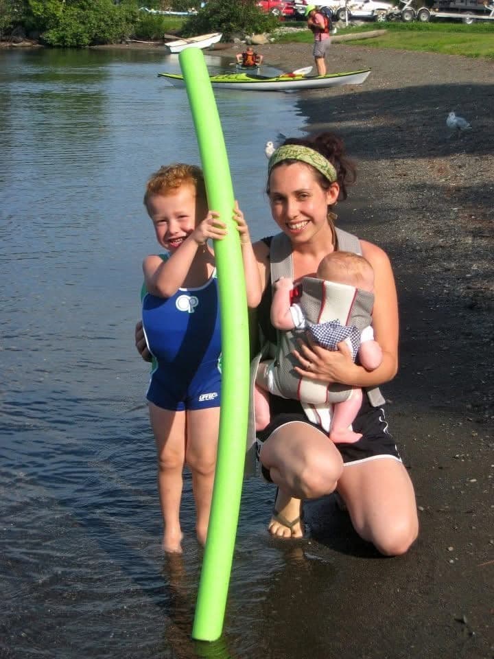 Oliver's family at the water's edge: a toddler holding a pool noodle in the shallows, and a baby in a carrier